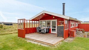 Een rode houten chalet met terras, schoorsteen en eethoek met stoelen en tafel.