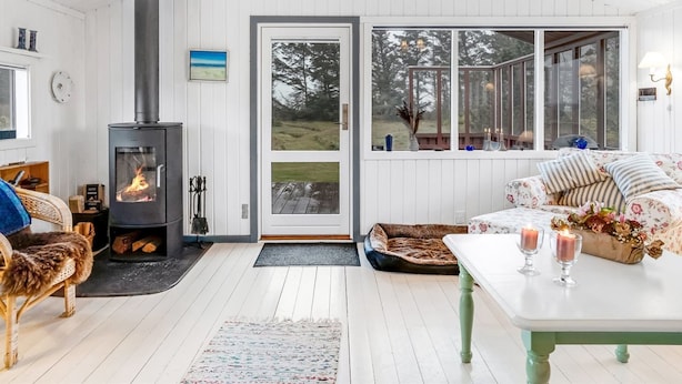 Een gezellige woonkamer met een houtkachel, een bank, een eettafel met kaarsen en een groot raam met uitzicht op de natuur.