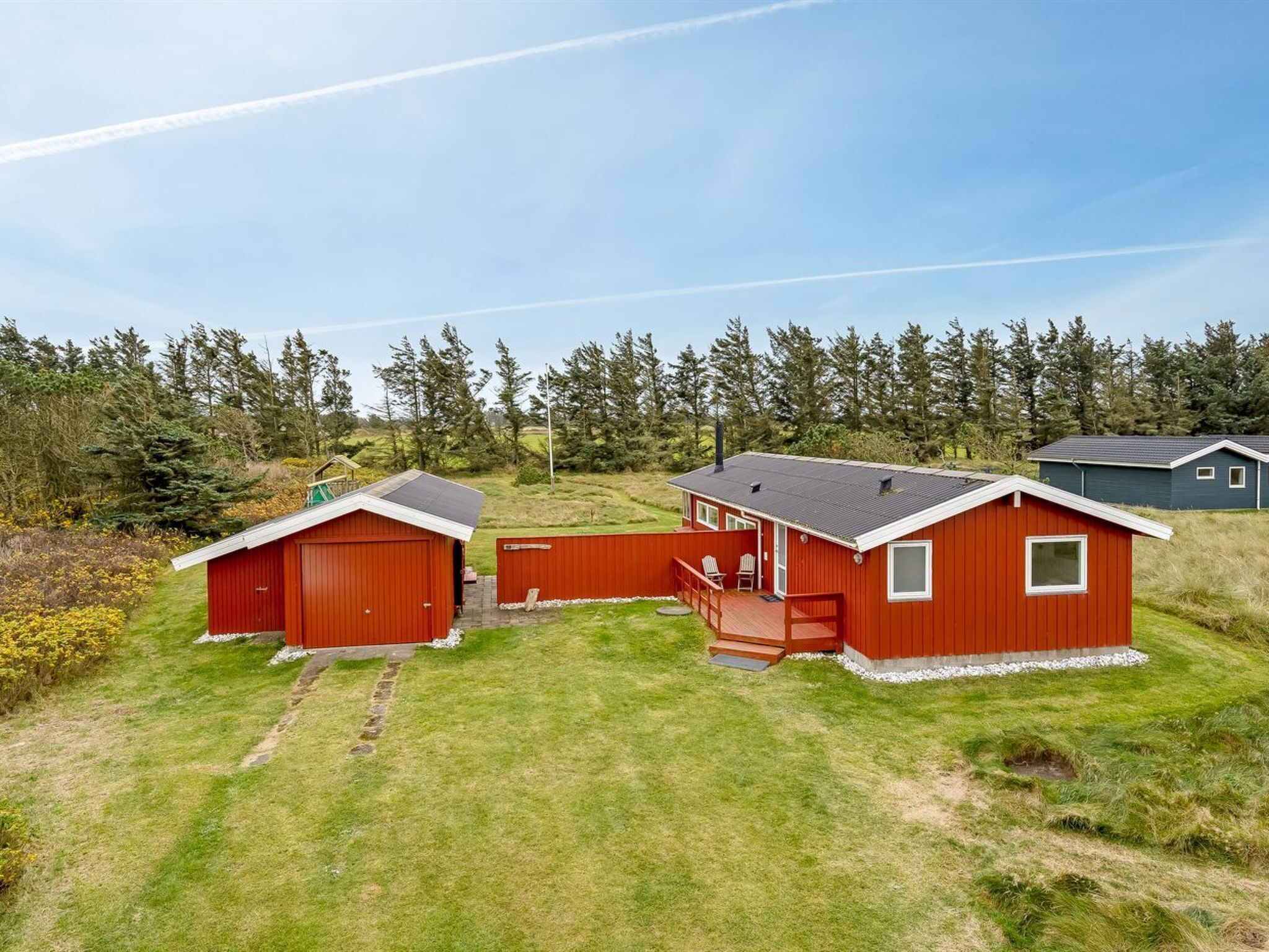 Une cabane rouge avec un porche et un garage, entourée d’une pelouse et d’arbres.