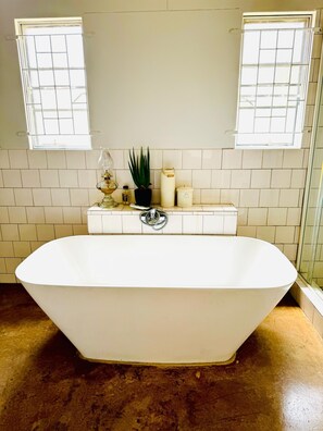 A large freestanding bathtub in a bathroom with white tiled walls, a potted plant, and a window with a grid pattern.