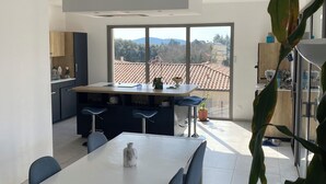 A modern kitchen with a large island, a dining area with a white table and blue chairs, and a view of a tiled roof and trees through large windows.