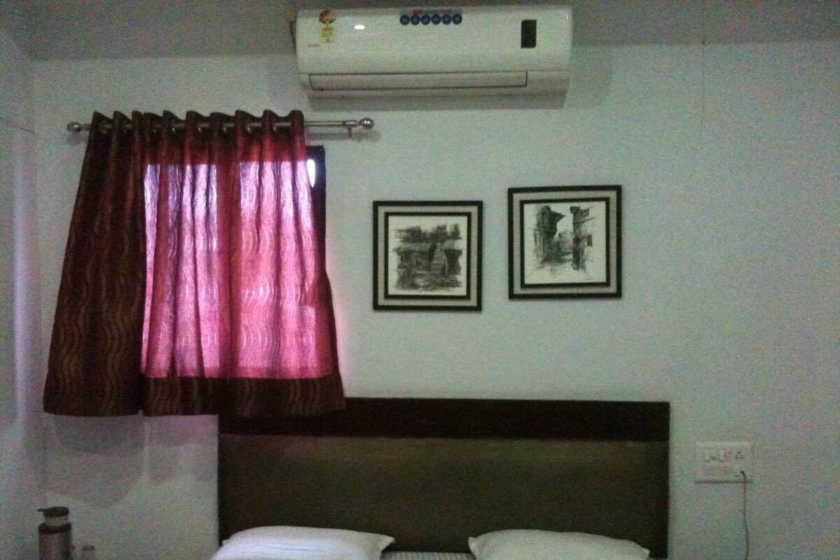 A bedroom with a bed, a nightstand, a lamp, a wall-mounted air conditioner, and two framed pictures.