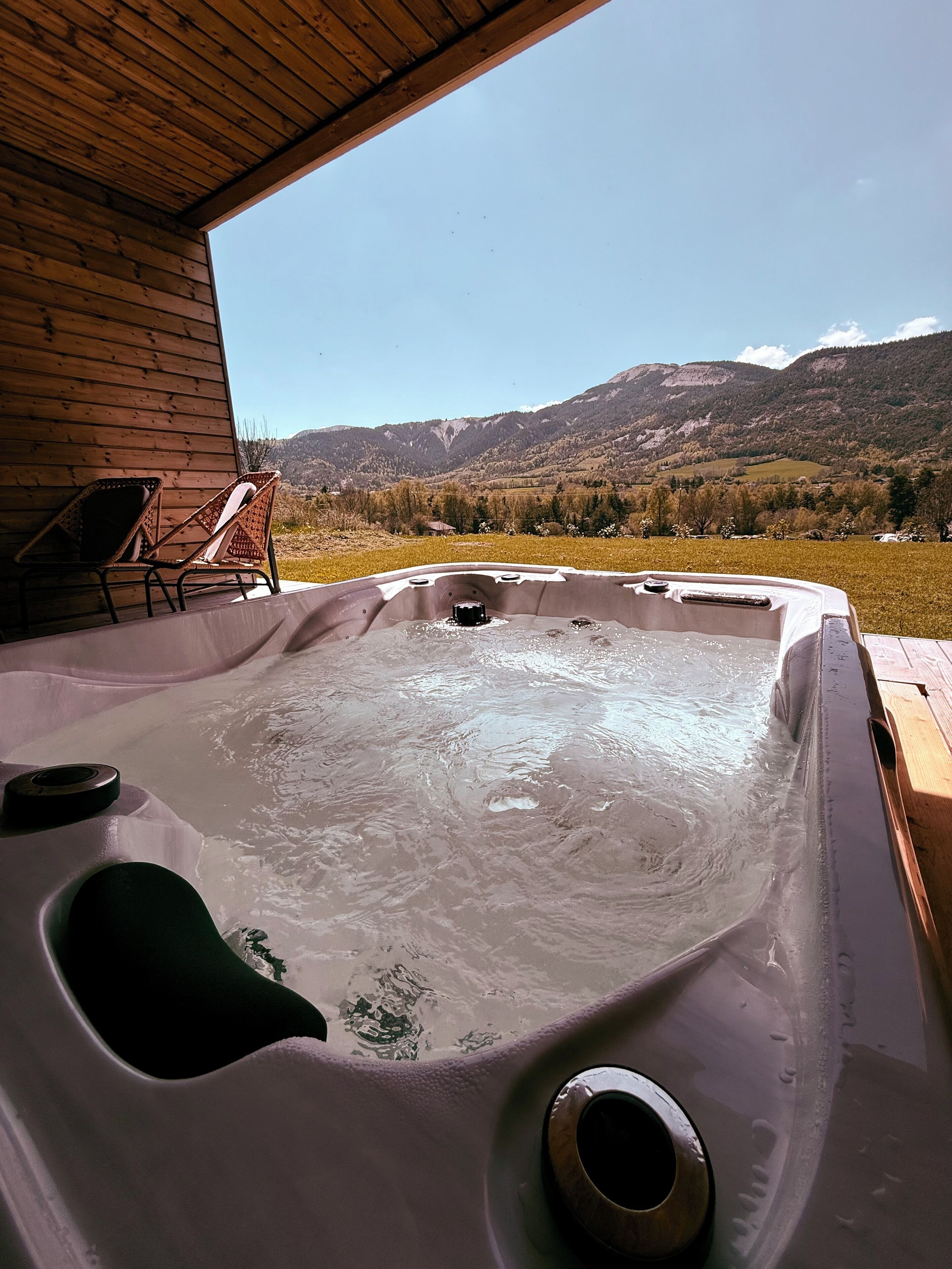 A hot tub with a wooden deck and chairs, overlooking a mountainous landscape.