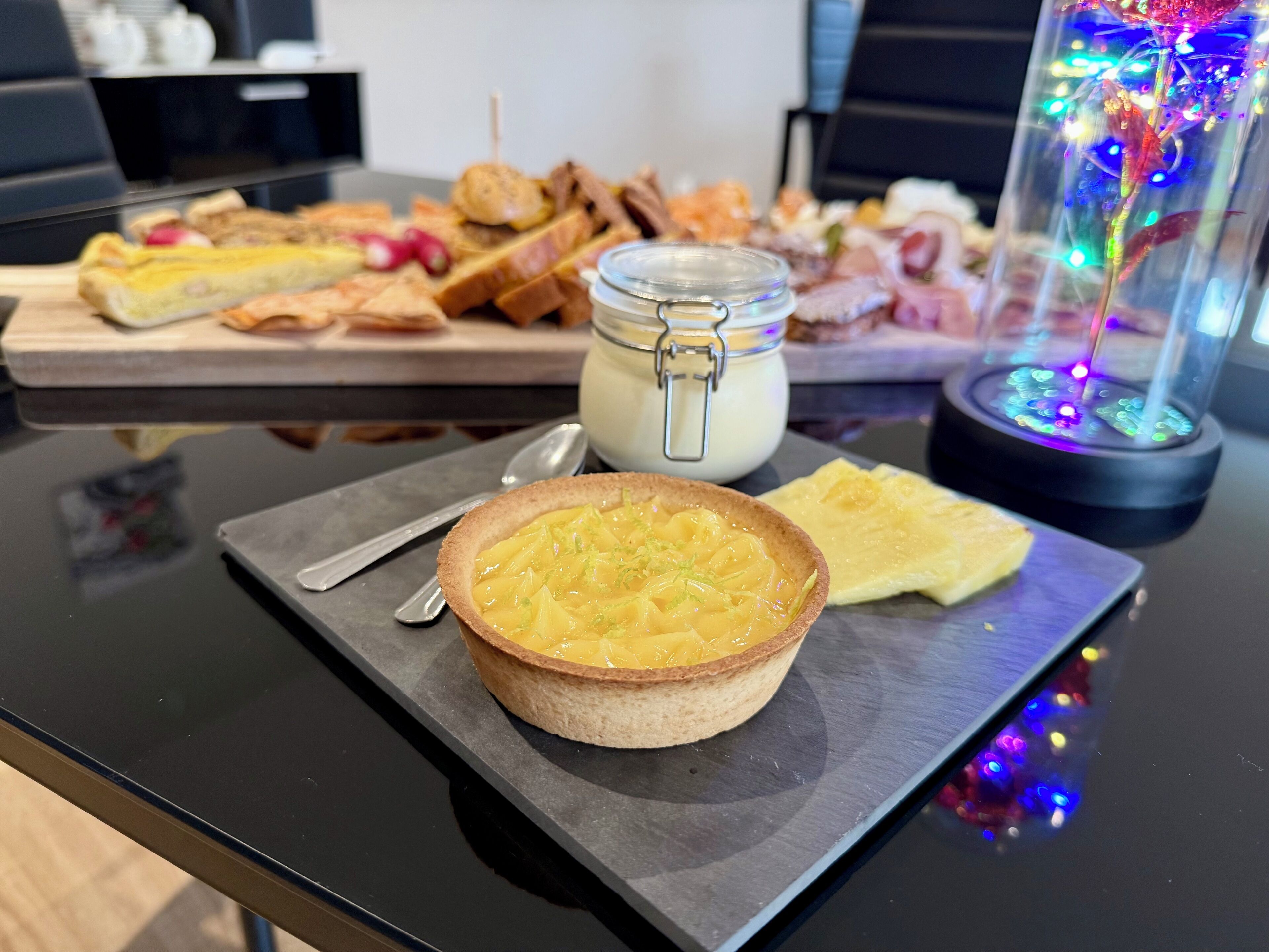 Un plato de postre con una tarta, una porción de pastel, un tarro de crema y un vaso decorativo con luces.