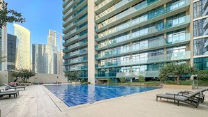A modern building with a swimming pool, lounge chairs, and a clear view of surrounding high-rise buildings. - Dubai 1BR | Merano Canal View Retreat Near Metro Dining Shops Leisure (Business Bay)