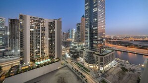 A high-rise building with multiple floors, surrounded by other tall buildings, a river, and a busy road. - Dubai 1BR | Merano Canal View Retreat Near Metro Dining Shops Leisure (Business Bay)