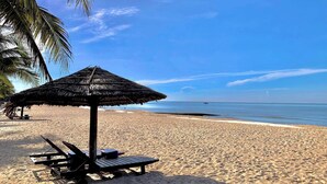 A beach with a thatched umbrella and lounge chairs.