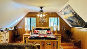 A cozy attic bedroom with a sloped ceiling, wooden walls, a bed with patterned bedding, two bedside lamps, a ceiling fan, and a large landscape painting. - Rustic Sierra Cabin with Hiking Trail and Kids Room - Perfect Family Getaway! (Mi-Wuk Village)