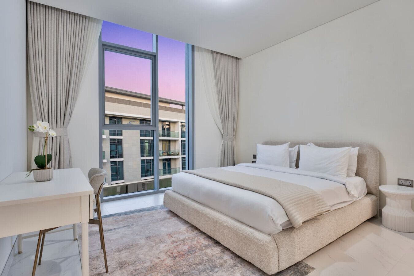 A modern hotel room with a large bed, a desk, and a view of a building through floor-to-ceiling windows.