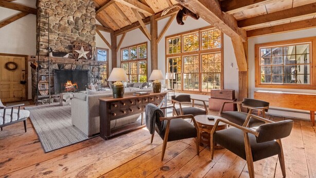 Ein rustikaler Innenraum mit Kamin aus Stein, Holzbalken und großen Fenstern.