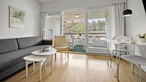 Un salon moderne avec un canapé gris, une petite table ronde, une chaise en bois et une table à manger avec des chaises. Une grande fenêtre offre une vue sur les arbres et un immeuble.