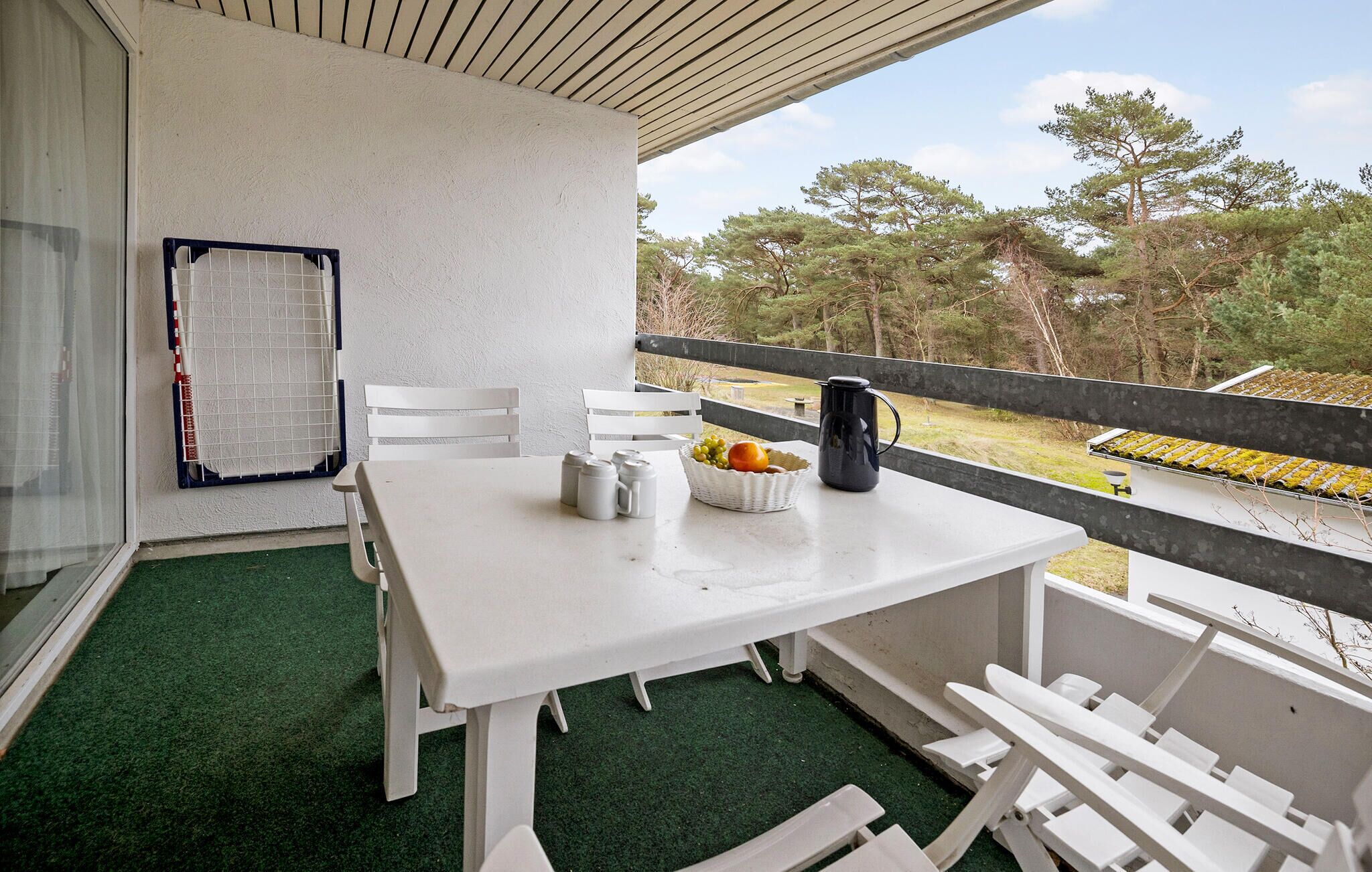 Ein Balkon mit einem weißen Tisch, Stühlen, einer Obstschale, Salz- und Pfefferstreuern und einer schwarzen Kaffeekanne.