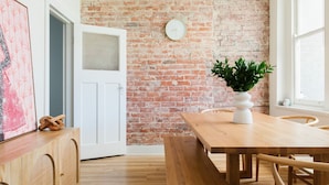 Una habitación con pared de ladrillo, una mesa de madera, un banco, una puerta blanca y una planta en maceta.