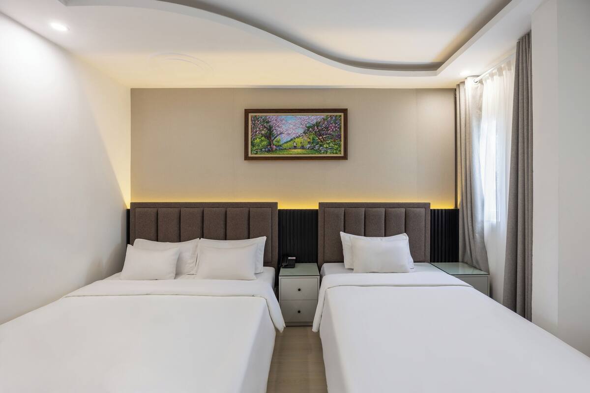 Ein Hotelzimmer mit zwei Betten, einem Nachttisch mit Lampe, einem Bild an der Wand und einem Fenster mit Vorhängen.