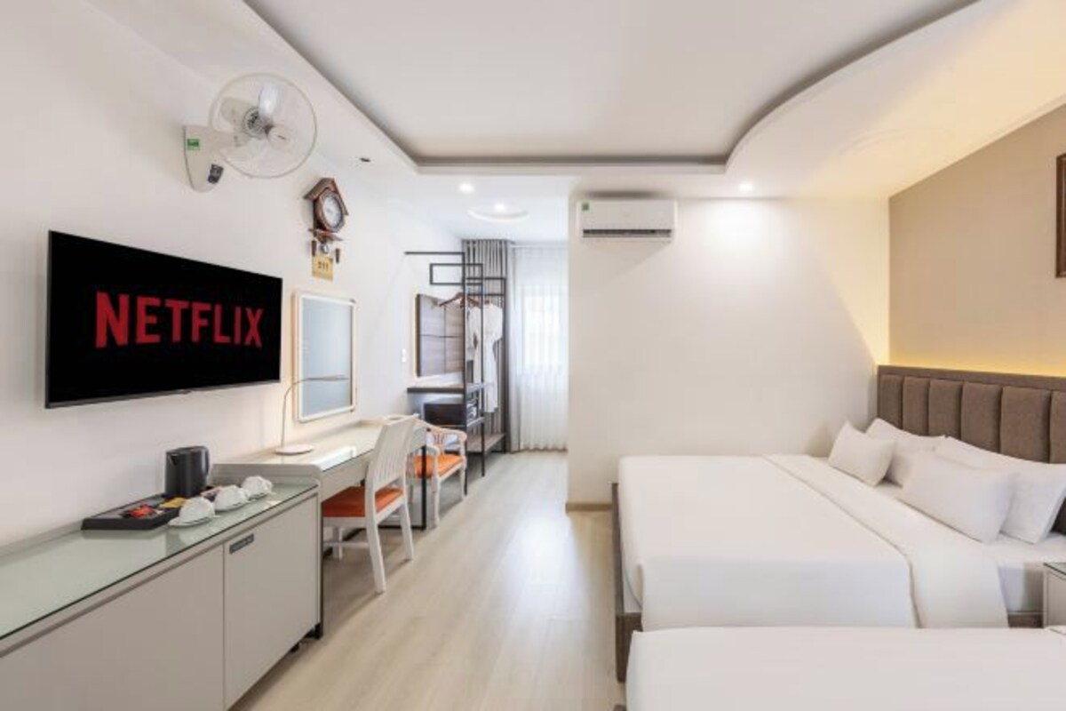 A modern hotel room with a large flat-screen TV, a desk with a chair, two beds, a wardrobe, and a ceiling fan.