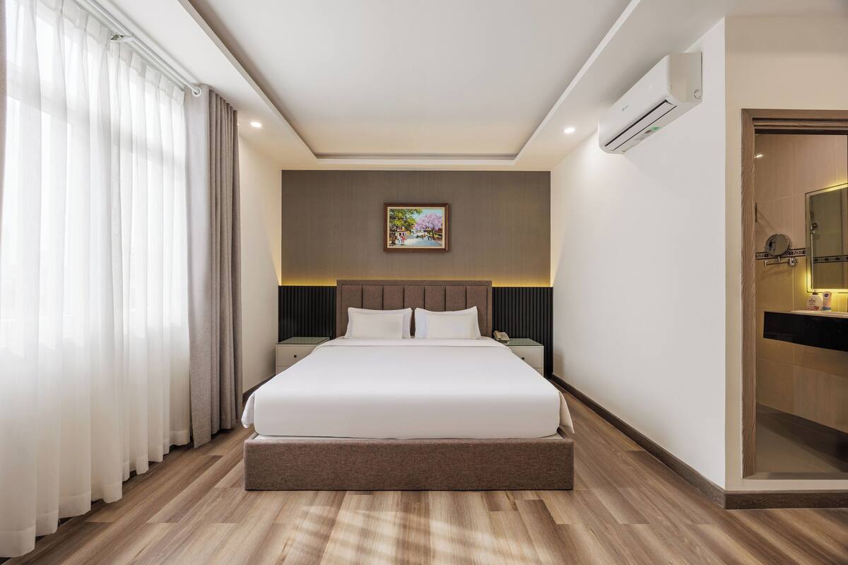 Ein Hotelzimmer mit einem großen Bett, einem Nachttisch, einem kleinen Tisch daneben, einem Bild an der Wand und einer Klimaanlage.