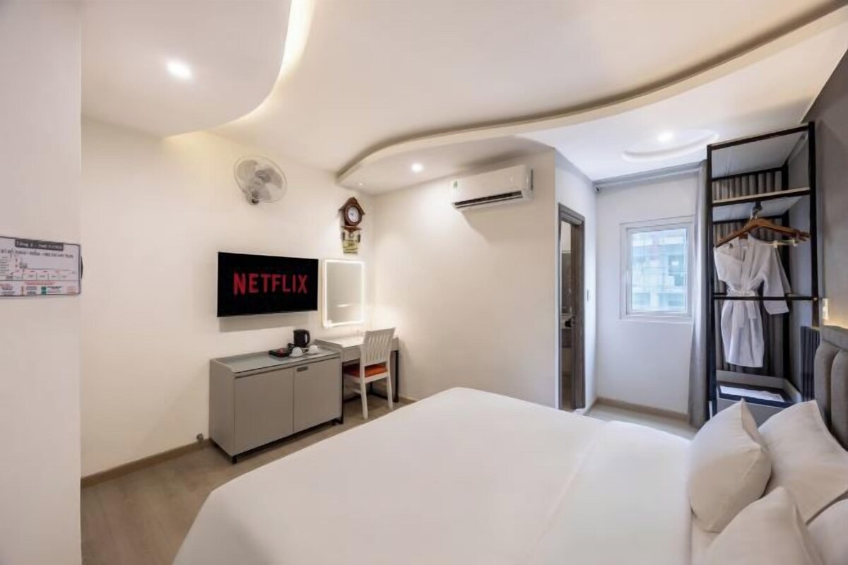 A modern hotel room with a bed, a desk with a chair, a wardrobe, a TV displaying 'NETFLIX', and a window.
