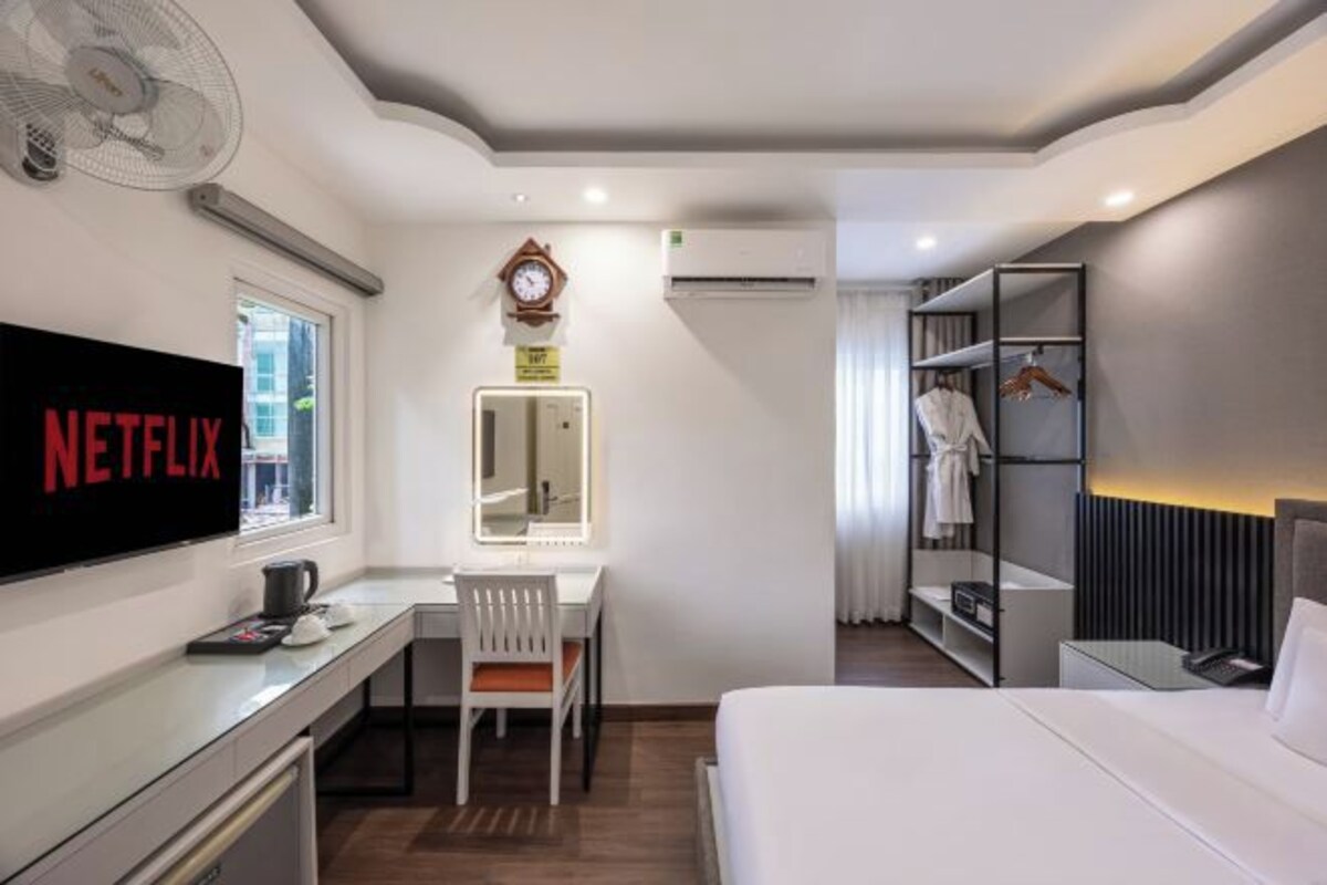 A modern hotel room with a bed, a desk with a chair, a TV, a mirror, a wardrobe, and a fan.