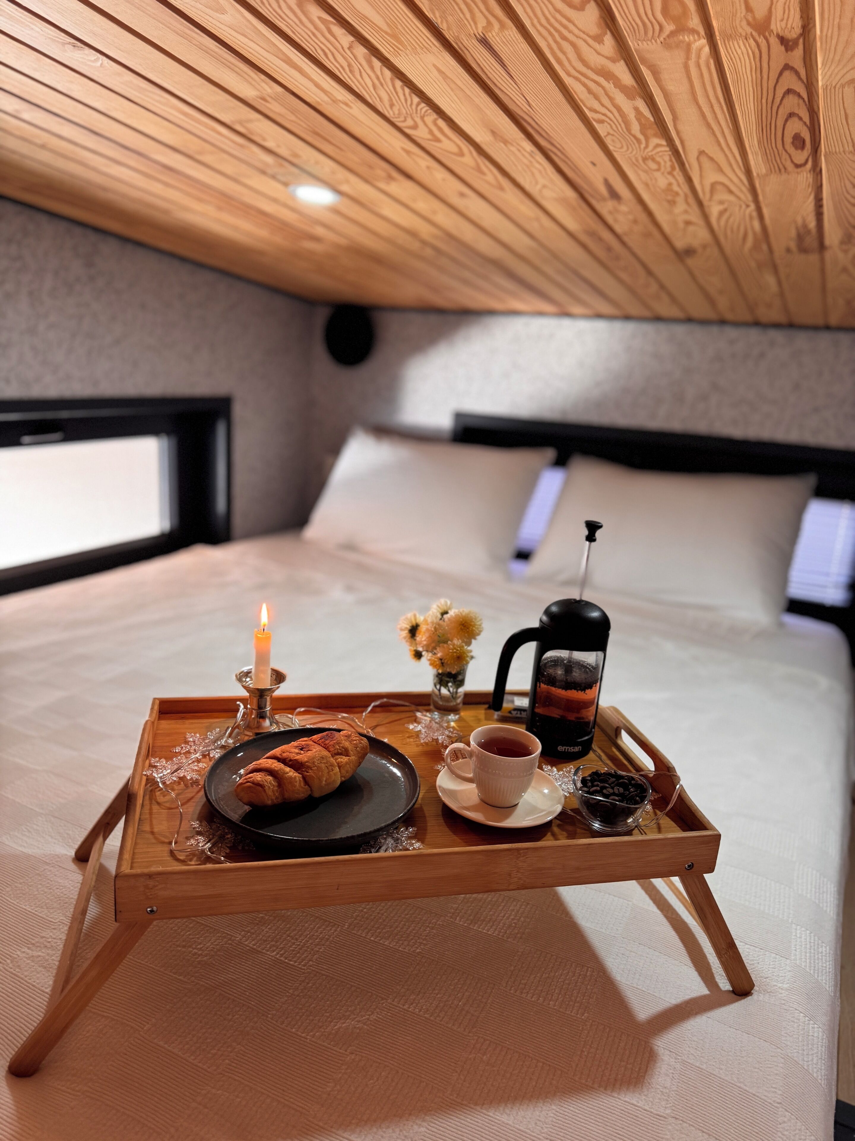 Ein gemütliches Schlafzimmer mit Holzboden, einem Bett mit weißen Leinen, einem Nachttisch mit Tablett, auf dem ein Croissant, eine Tasse Tee und eine Kaffeemaschine stehen.
