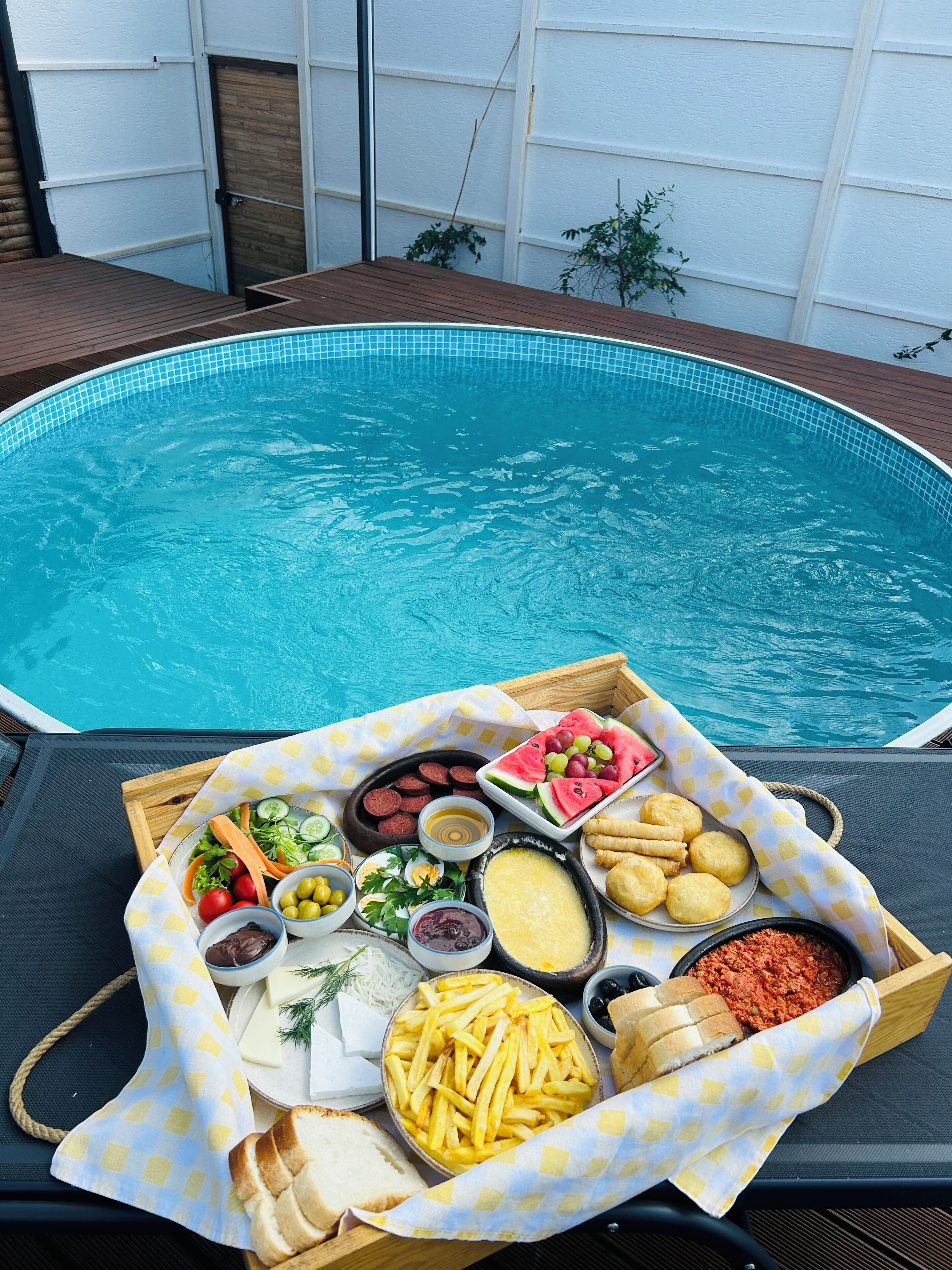 Ein Pool-Buffet mit verschiedenen Speisen und Getränken, darunter Würstchen, Käse und Brot, mit Blick auf das Schwimmbecken.