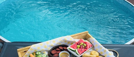 A poolside buffet with various foods and drinks, including sausages, cheese, and bread, set against a pool backdrop.