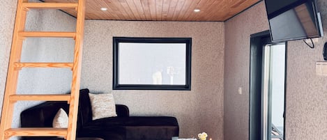 A cozy room with a loft bed, a wooden ladder, a dark sofa, a small table with a vase of flowers, and a window with a view.