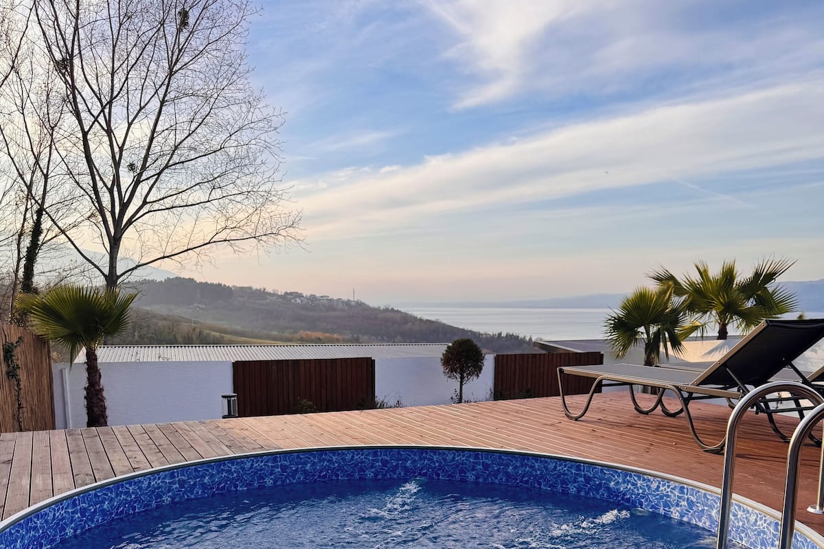 Piscina con un bol de fruta tropical y copas de vino en una terraza de madera, con vistas a un cuerpo de agua y colinas.