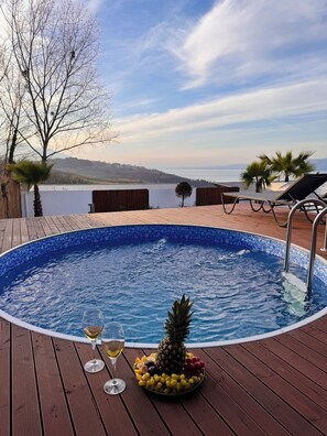 Une piscine, un saladier rempli de fruits tropicaux et des verres à vin disposés sur une terrasse en bois, le tout offrant une vue sur une étendue d’eau et des collines.