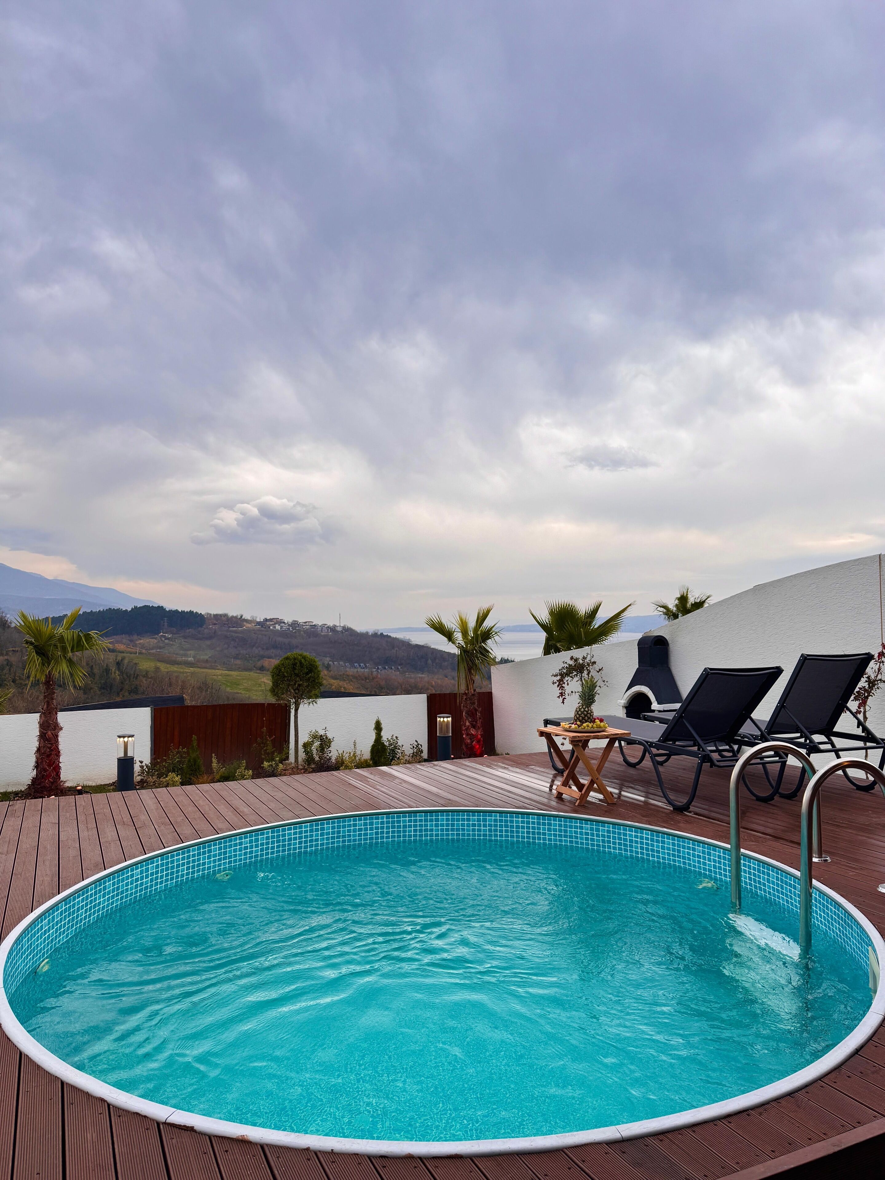 A circular pool with a wooden deck, two lounge chairs, and a table with fruits.