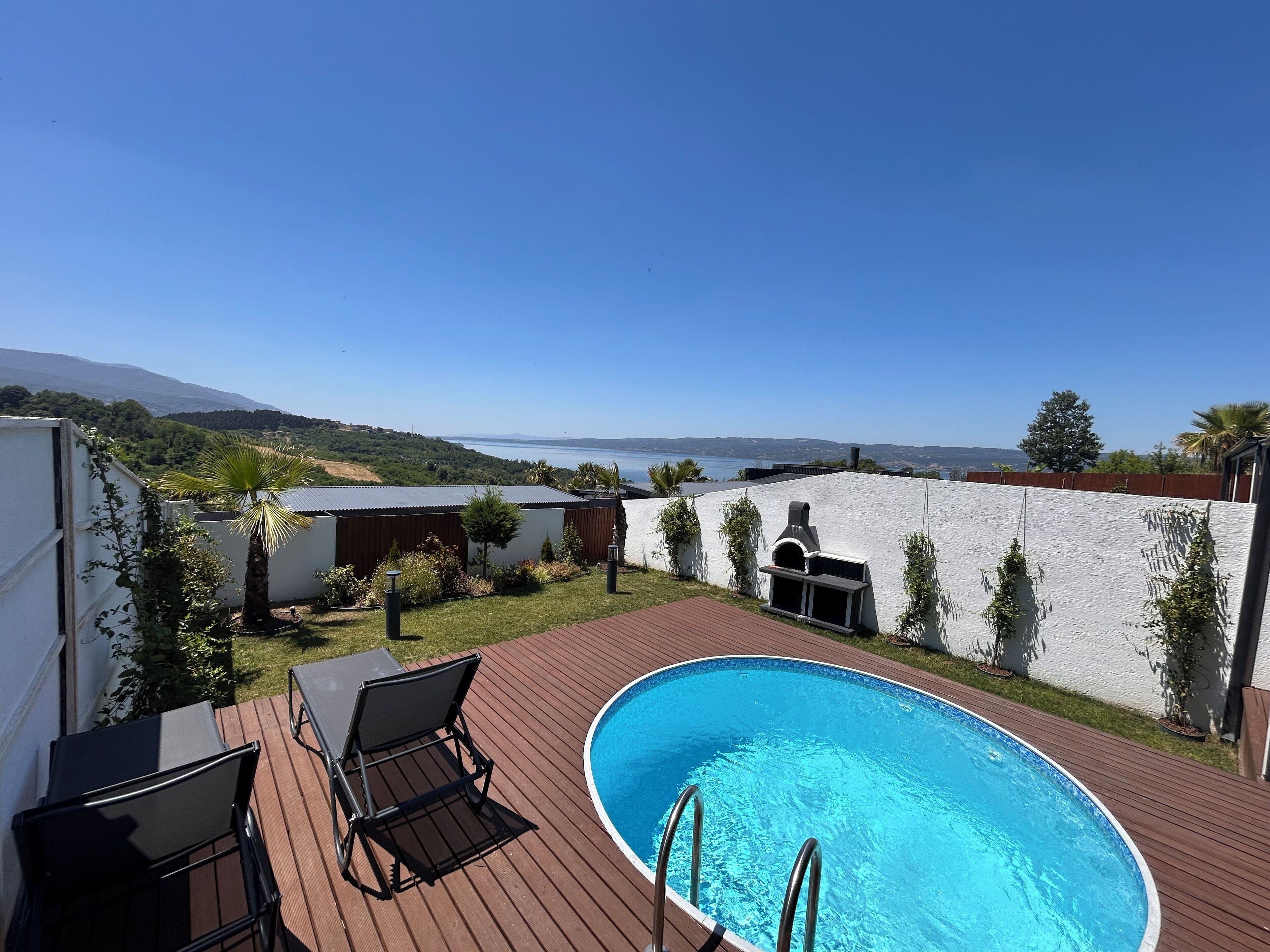 A swimming pool with a wooden deck, surrounded by a white wall and a view of the sea and hills.