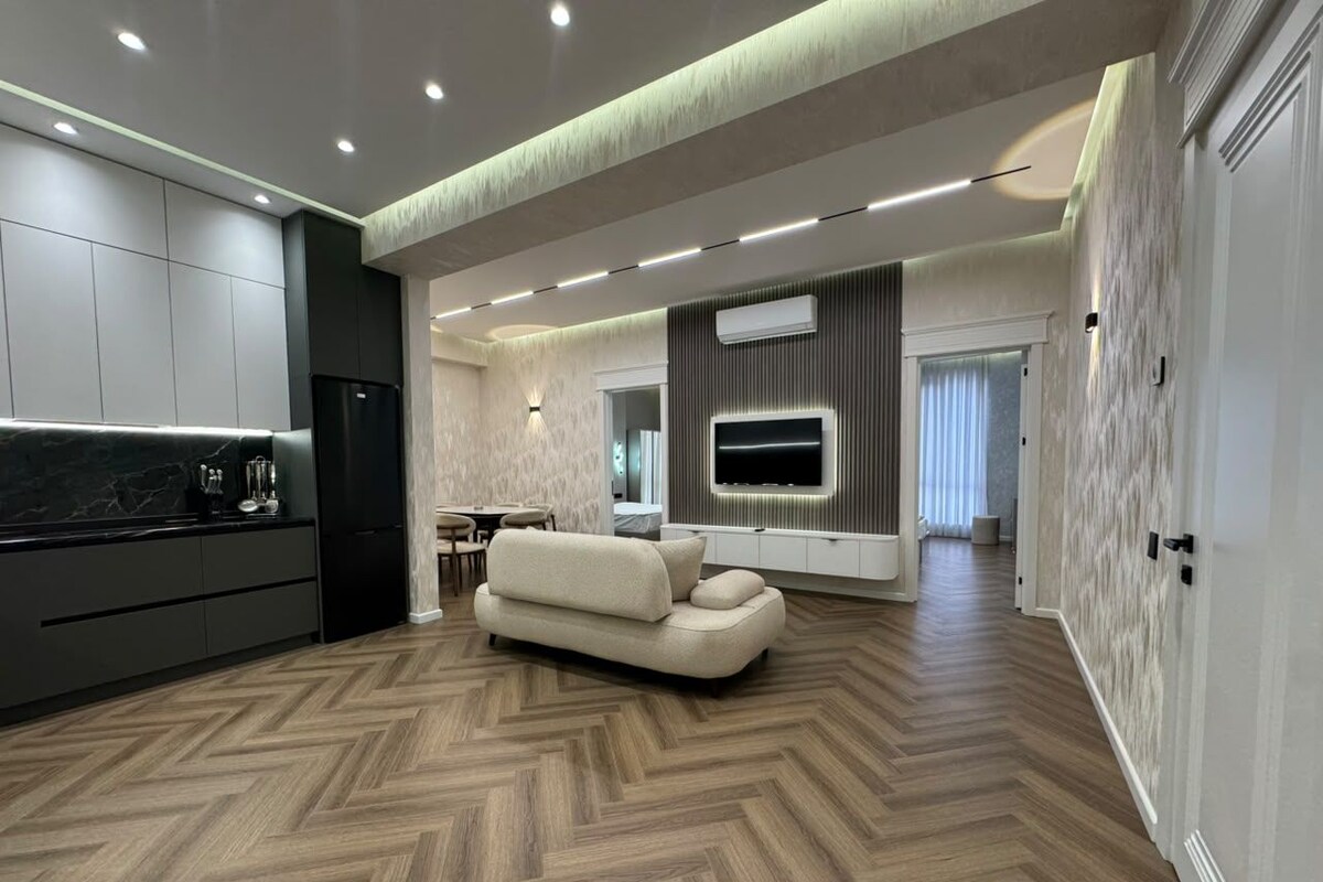 A modern living room with a herringbone floor, a sofa, a TV mounted on the wall, and a kitchen area with cabinets and a refrigerator.