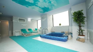 Ett modernt inomhusbadområde med en blå mosaikbeklädd pool, två solstolar, en krukväxt och ett tak med ett molnmotiv.