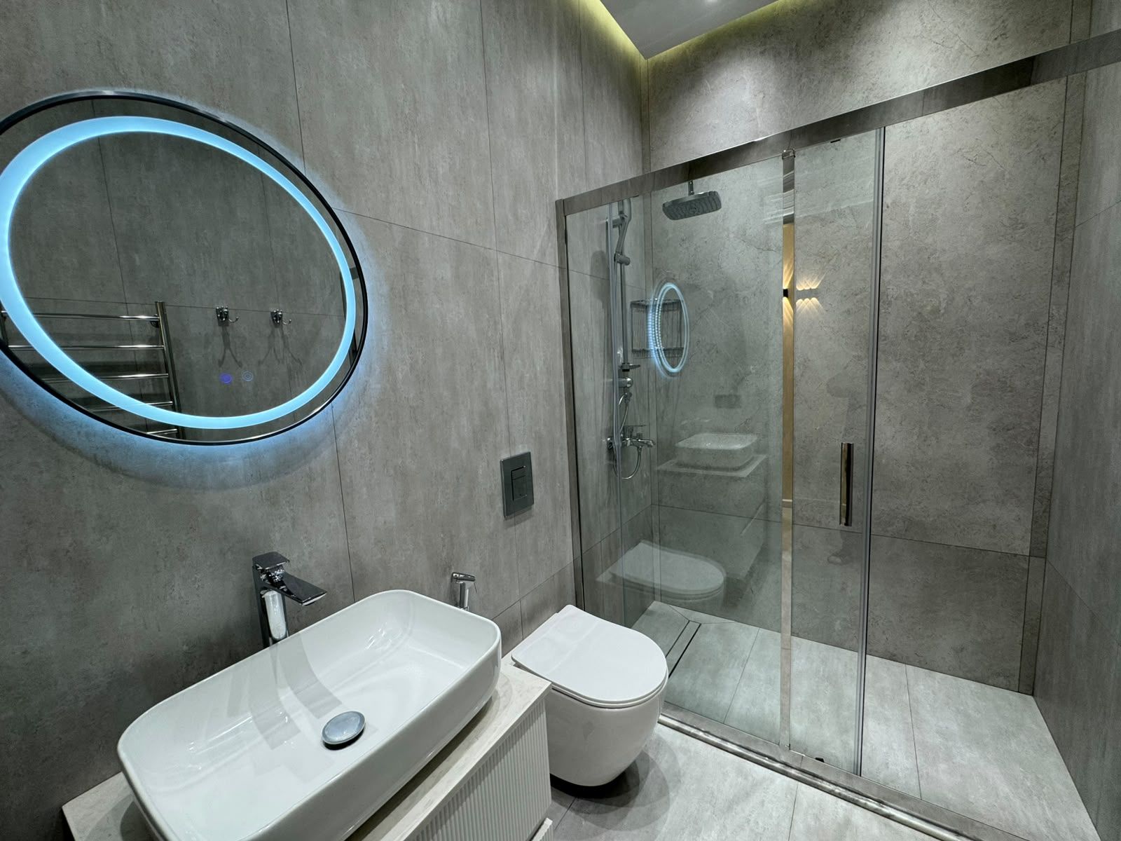 Un baño moderno con ducha envuelta en vidrio, un espejo redondo con iluminación LED, un lavamanos blanco y un inodoro.