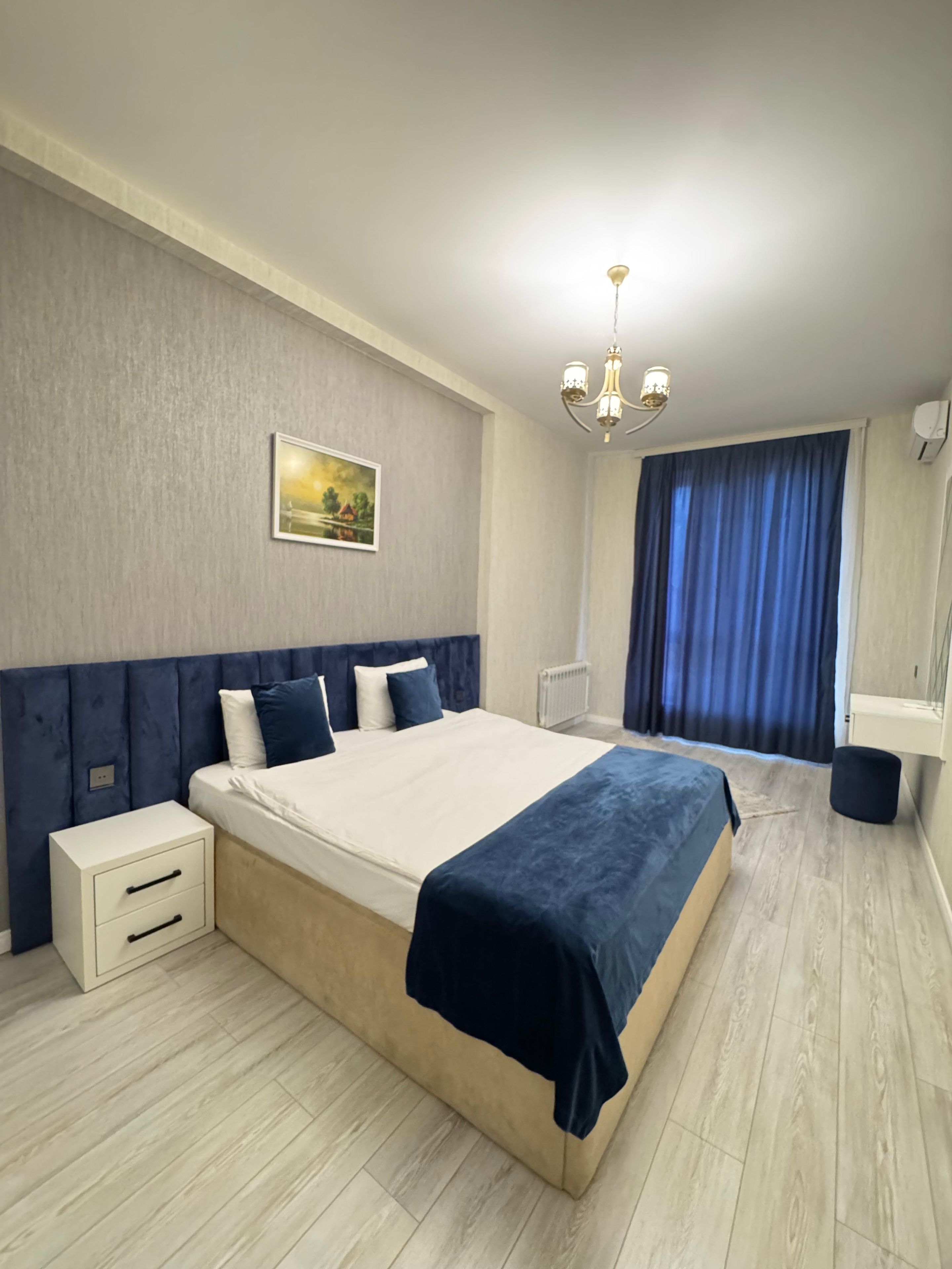 A modern bedroom with a large bed, bedside tables, a nightstand, a painting on the wall, a chandelier, and a window with curtains.