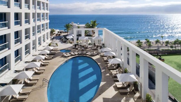 A hotel pool area with multiple sun loungers and umbrellas, overlooking a beach and ocean.