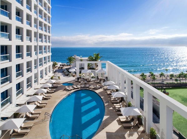 A hotel with a pool, lounge chairs, umbrellas, and a view of the ocean.
