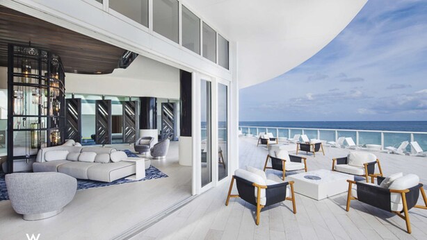 Ein modernes Interieur mit einer großen Couch, Sesseln und einem Couchtisch. Durch eine Glaswand bietet sich Meerblick.
