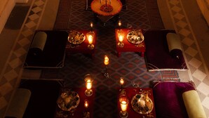 Una habitación ricamente decorada y tenuemente iluminada, con una mesa central dispuesta para cenar, rodeada de asientos.