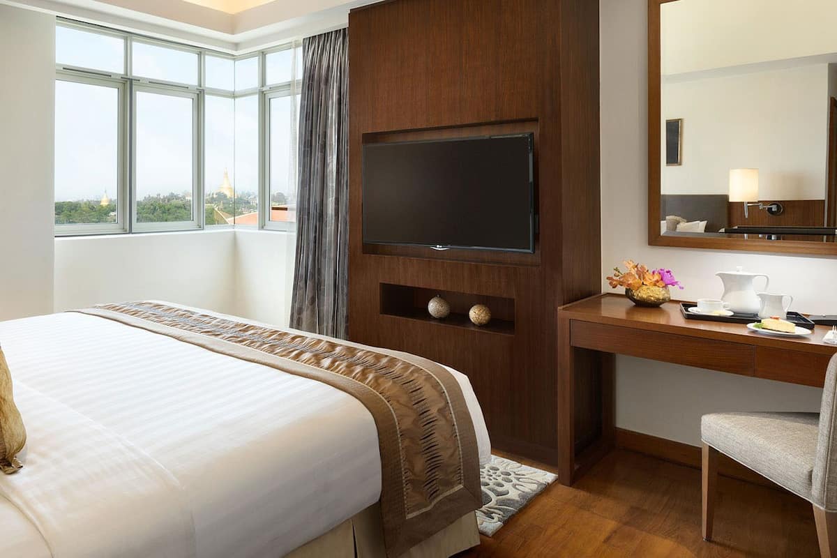Ein Hotelzimmer mit einem großen Bett, einem Schreibtisch, einem Flachbildfernseher, einem Sessel und einem Fenster mit Blick auf eine Stadtlandschaft.