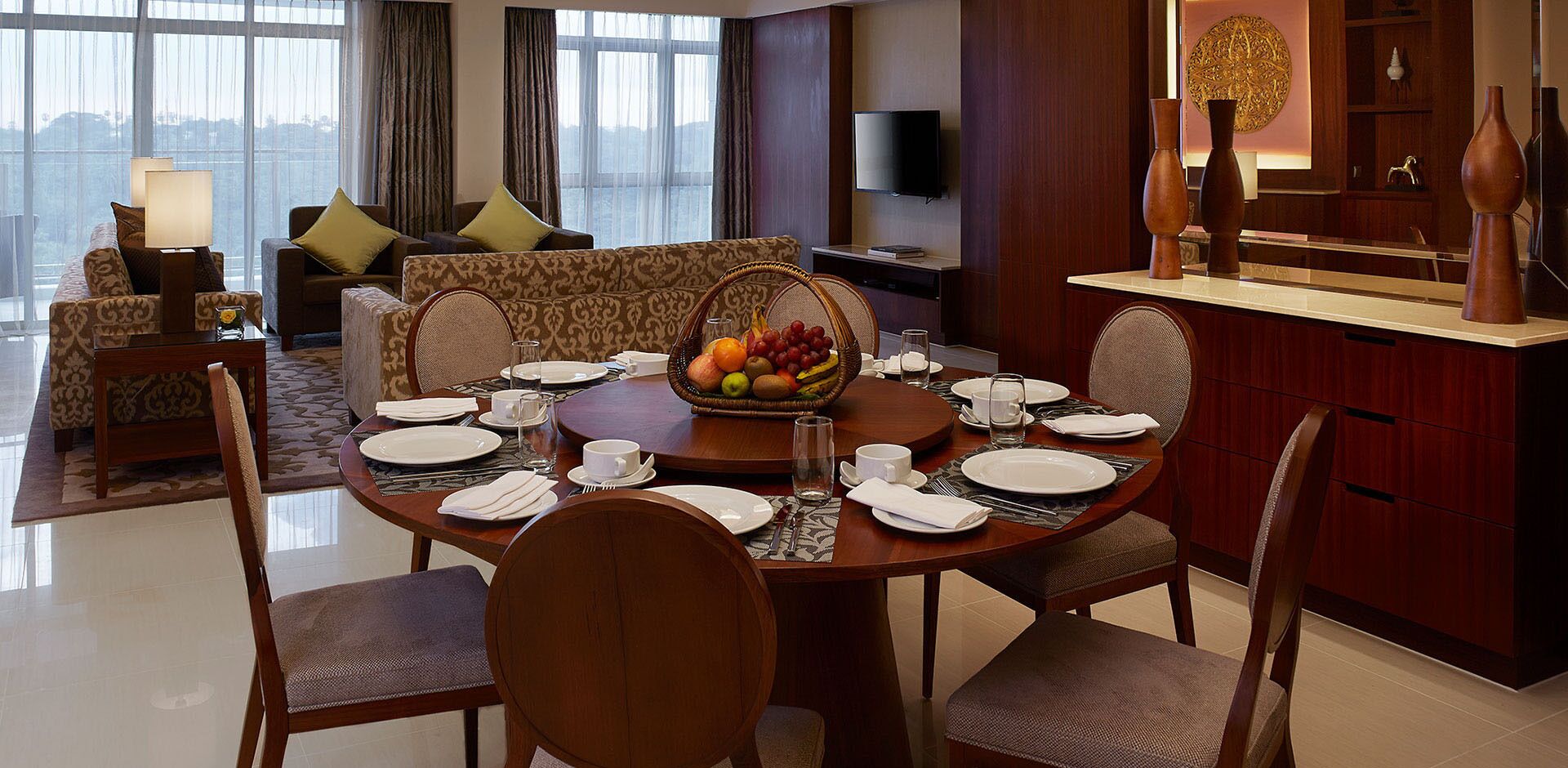 A dining area with a round table set for four, surrounded by chairs. A fruit basket is placed on the table, and there is a television mounted on the wall above the seating area.