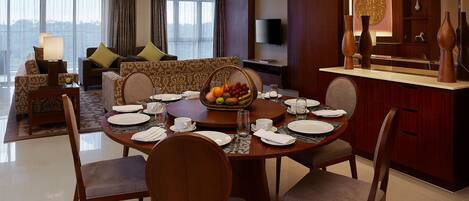 A dining area with a round table set for four, surrounded by chairs. A fruit basket is placed on the table, and there is a television mounted on the wall above the seating area.