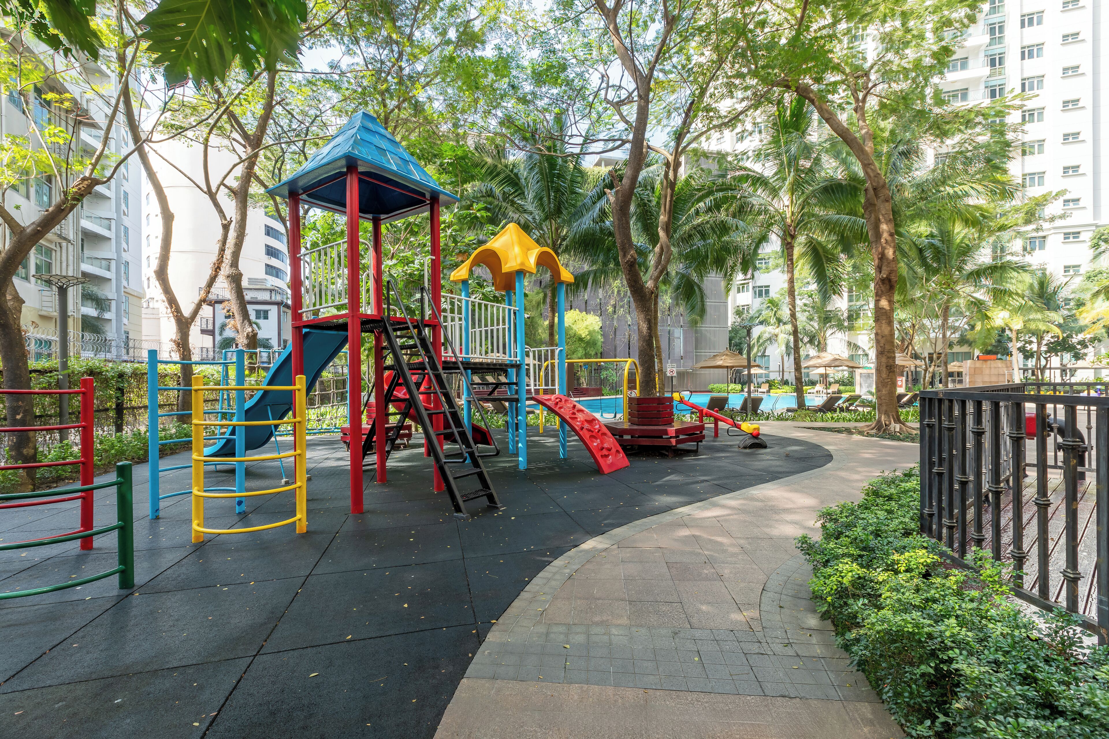 A playground with a slide and climbing structures, surrounded by trees and a paved path.