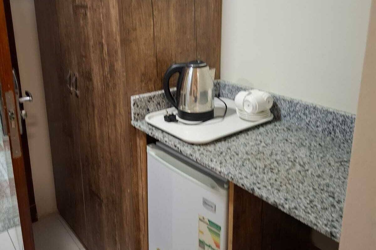 A compact kitchen with a wooden door, a small refrigerator, and a countertop with a kettle and a white appliance.