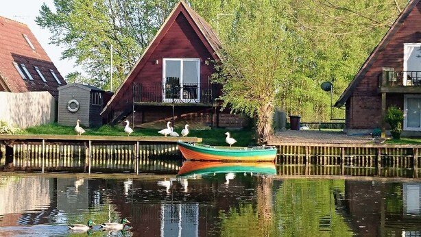 A row of wooden houses with a canal in front, a colorful boat, and birds on the dock. - River Lark Lodge (Isleham)