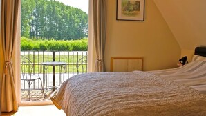 A bedroom with a large bed, a small table, and a view of a green landscape through a balcony. - River Lark Lodge (Isleham)