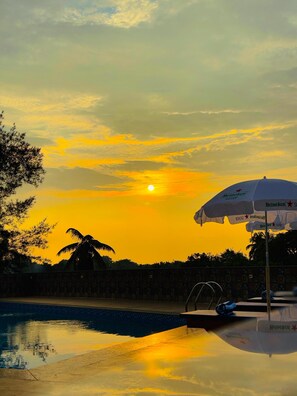 A sunset over a swimming pool with a Heineken umbrella and palm trees. - Nazri Hotel (Arambol)