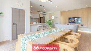 A modern living room with a dining area, a kitchen in the background, and a TV mounted on the wall.