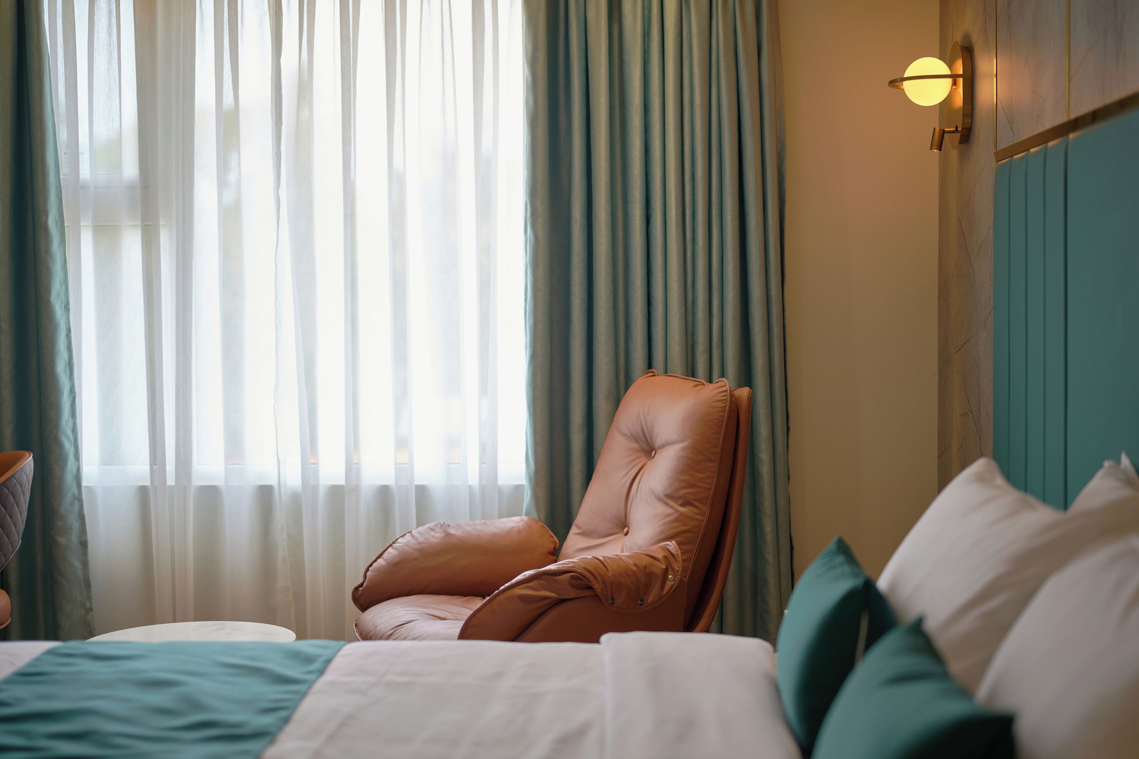 Een hotelkamer met een bed, een leren fauteuil en een raam met dunne gordijnen.