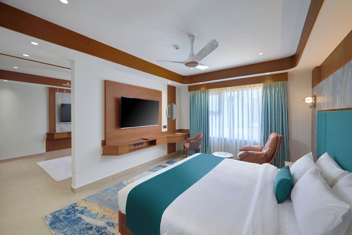 A modern hotel room with a large bed, a flat-screen TV mounted on the wall, a desk with a chair, and a ceiling fan.