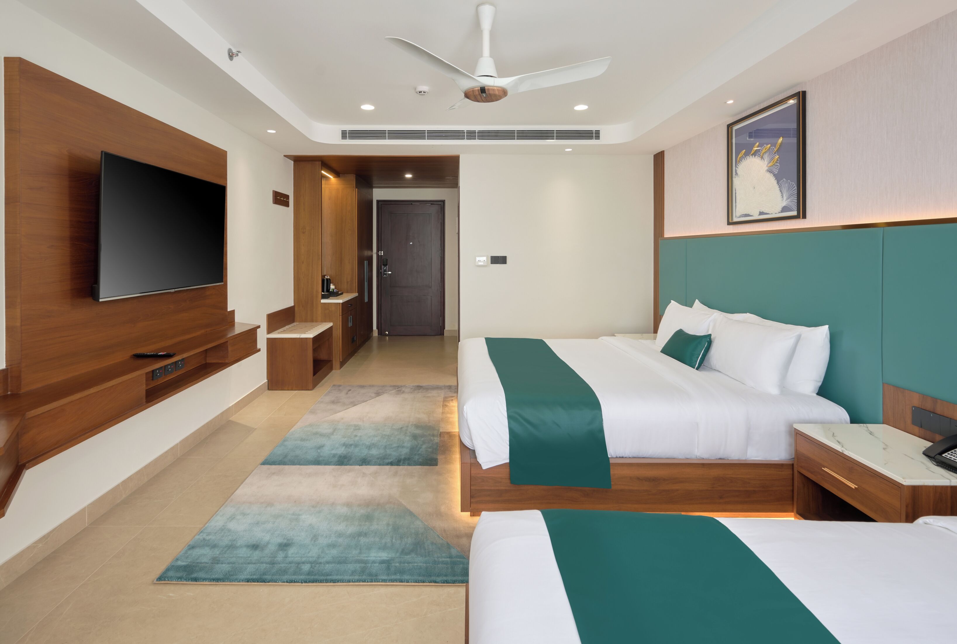 A modern hotel room with a large flat-screen TV, two beds with green bedding, a ceiling fan, and a wooden headboard.